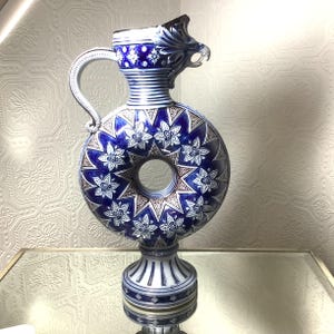 May include: A blue and white ceramic pitcher with a ring-shaped body and a dragon head spout. The pitcher has a detailed floral design and stands on a pedestal base.