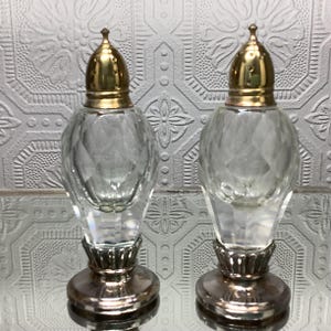 Puede incluir: Dos saleros y pimenteros de vidrio transparente con lados facetados y tapas de metal dorado. Los saleros y pimenteros tienen una base de metal plateado y se exhiben sobre una superficie reflectante.