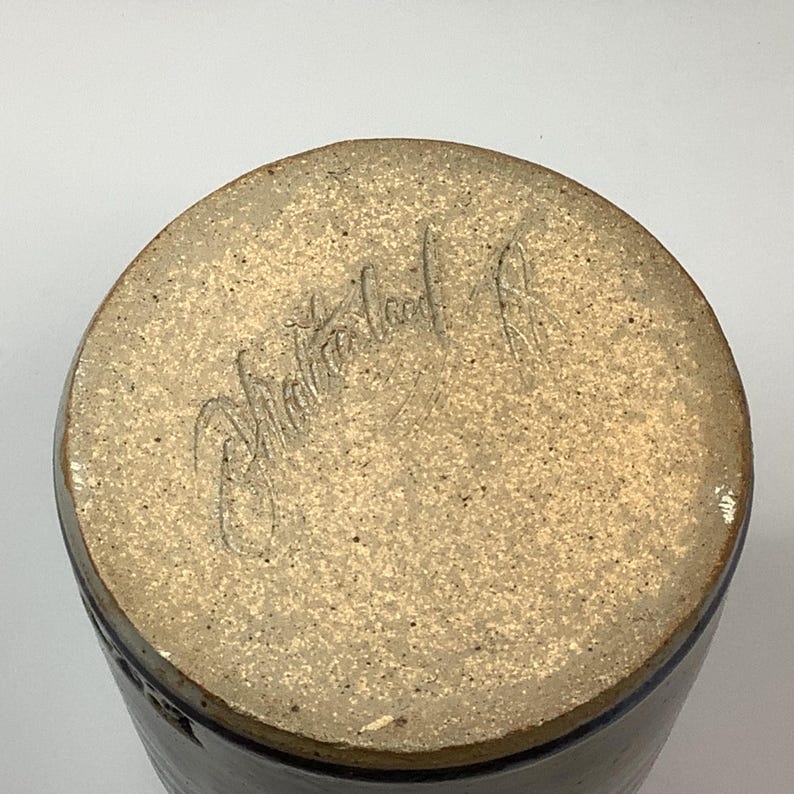 Puede incluir: La base circular de un recipiente de cer&aacute;mica con una superficie de arcilla beige moteada y sin esmaltar. Una firma manuscrita en cursiva est&aacute; grabada en el centro de la base, con el n&uacute;mero 98 visible a la derecha de la escritura.