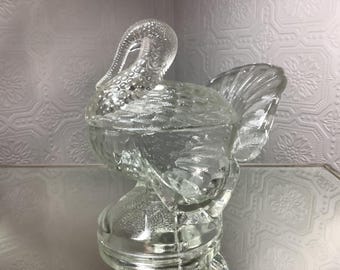 May include: A clear glass turkey-shaped candy dish with a lid. The dish features a detailed design, including a textured body, tail feathers, and a curved neck. The glass is transparent, allowing light to pass through.