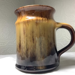 Op de afbeelding: Een keramische mok met een bruine en bruingele glazuur. De mok heeft een afgeronde body, een oor en een licht uitlopende rand. De kleuren vloeien in elkaar over, waardoor een rustieke, aardse uitstraling ontstaat. De mok is ongeveer 13 cm hoog.