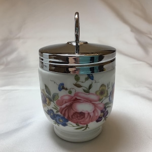 May include: A white ceramic egg cup with a floral design and a silver lid with a handle.