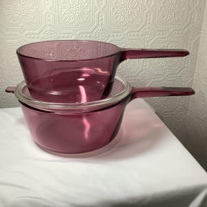 May include: Two vintage, ruby-colored glass saucepans with long handles. One saucepan is stacked on top of the other. The bottom saucepan has a clear glass lid. The saucepans are on a white surface.