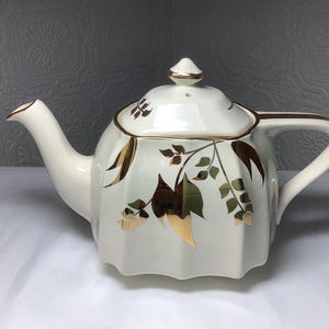 May include: A white ceramic teapot with a lid, spout, and handle. The teapot is decorated with gold trim and floral designs in gold, brown, and green. The teapot is sitting on a white surface.