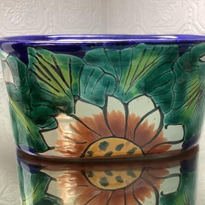May include: An oval ceramic planter featuring a hand-painted design of large green leaves and a central orange sunflower with a yellow centre. The rim is a solid deep blue, and the base of the planter is white with black outlines.