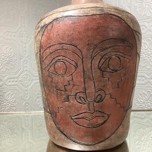 Peut inclure: Un vase en céramique avec un motif de visage. Le vase est principalement brun rougeâtre avec un bord et une base beige clair. Le visage est souligné en noir, avec de grands yeux, un nez et une bouche. Le design a un style abstrait et artistique.