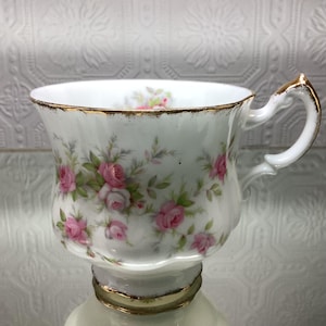 May include: White teacup with a floral rose pattern in pink and green. The cup has a gold rim and handle. The teacup is sitting on a white saucer with a gold rim.