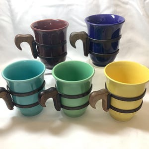 Puede incluir: Cinco tazas de cerámica con asas de madera. Las tazas son de diferentes colores: azul, marrón, verde, turquesa y amarillo. Cada taza tiene una banda negra alrededor del medio.