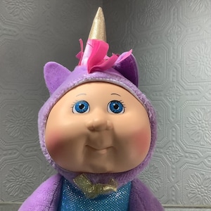 May include: A Cabbage Patch Kids doll wearing a purple unicorn costume with a gold horn and pink accents. The doll has blue eyes and rosy cheeks. The costume includes a blue sequined top and a gold bow.