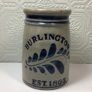Puede incluir: Una vasija de gres cil&iacute;ndrica con acabado beige moteado y borde azul oscuro. El frente presenta el texto azul en relieve BURLINGTON sobre un motivo de hoja azul y el texto EST. 1865 en la base. El recipiente tiene un borde superior grueso.