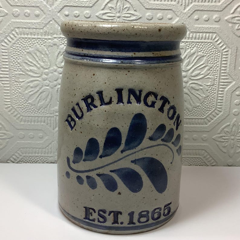 Puede incluir: Una vasija de gres cil&iacute;ndrica con acabado beige moteado y borde azul oscuro. El frente presenta el texto azul en relieve BURLINGTON sobre un motivo de hoja azul y el texto EST. 1865 en la base. El recipiente tiene un borde superior grueso.