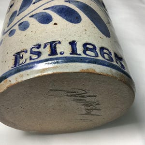 Puede incluir: Un recipiente cil&iacute;ndrico de cer&aacute;mica con esmalte blanquecino moteado y patrones de hojas pintados en azul. El texto EST. 1865 est&aacute; grabado en azul en el lateral. La base sin esmaltar presenta una firma oscura escrita a mano en la arcilla.