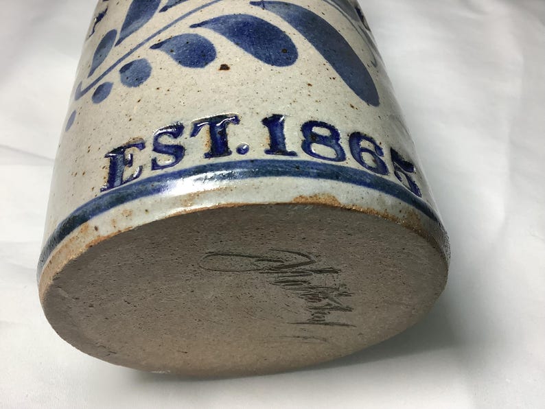 Puede incluir: Un recipiente cil&iacute;ndrico de cer&aacute;mica con esmalte blanquecino moteado y patrones de hojas pintados en azul. El texto EST. 1865 est&aacute; grabado en azul en el lateral. La base sin esmaltar presenta una firma oscura escrita a mano en la arcilla.