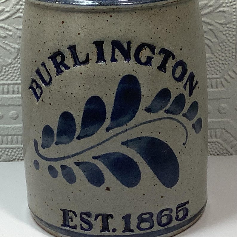 Puede incluir: Una vasija de gres cil&iacute;ndrica con acabado beige moteado y un dise&ntilde;o de hoja de helecho pintado a mano en azul oscuro. El texto BURLINGTON est&aacute; arqueado en la parte superior y el texto EST. 1865 est&aacute; impreso en azul oscuro en la base.