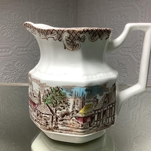 May include: White ceramic jug featuring a detailed village scene in brown, blue, and green. The rim and handle are white, with a brown patterned border. The jug has a hexagonal base and a curved handle.