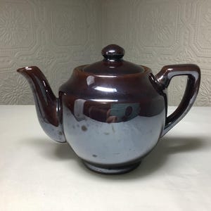 May include: A brown and blue ceramic teapot with a lid and handle. The teapot has a glossy finish and a rounded shape. The spout and handle are a darker brown than the body of the teapot. The teapot is sitting on a white surface.