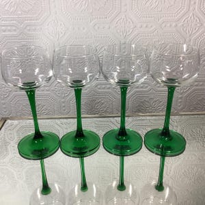 May include: Four clear wine glasses with green stems and bases. The glass bowls have a subtle etched design. The green stems and bases are a vibrant, solid color. The glasses are arranged in a row, showcasing their elegant design.