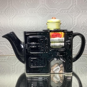 May include: A black ceramic teapot in the shape of a traditional stove. The teapot has a spout, handle, and a small yellow saucepan on top. The stove design includes oven doors and a firebox, with a stack of white and red items inside.