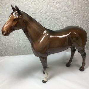 May include: A glossy ceramic horse figurine in shades of brown, with a white stripe on its face and white accents on its legs. The horse is posed in a standing position, showcasing its sleek, detailed design. A decorative piece for home decor.