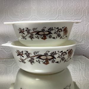 May include: Two off-white vintage Pyrex bowls with handles, stacked. Each bowl features a brown floral and vine pattern. The bowls are round with a slight rectangular shape and are reflected in the surface below.