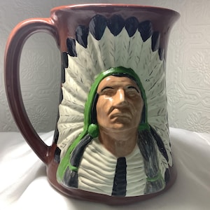 Op de afbeelding: Een keramische mok met een handvat, met een ontwerp van een Indiaanse chef. De mok heeft een bruine buitenkant, een witte en zwarte veren tooi en een groen en zwart kledingstuk. Het gezicht van de chef staat in het midden.