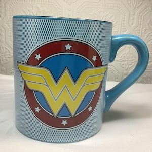 May include: A light blue ceramic mug with the Wonder Woman emblem. The emblem features a yellow 'W' with wings, set against a blue background within a red circle, and white stars. The mug has a light blue handle and a patterned exterior.