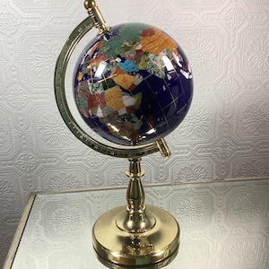 May include: A decorative globe with a blue ocean and colorful landmasses. The globe is supported by a gold-colored metal stand with a circular base and a curved meridian. The globe is a decorative item.