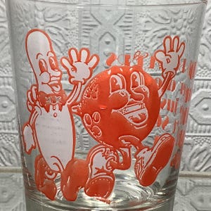 Puede incluir: Vaso de vidrio transparente con personajes de dibujos animados en naranja y blanco. Un personaje es un perrito caliente antropomórfico sonriente con pajarita. El otro es un personaje naranja sonriente con ojos grandes y una amplia sonrisa. El vaso tiene un patrón texturizado.