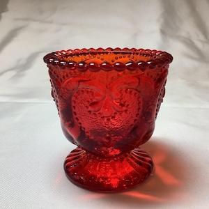 May include: A red glass candle holder with a beaded rim and a heart-shaped design. The holder has a pedestal base.