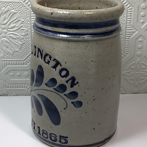 Puede incluir: Una vasija de gres cil&iacute;ndrica con acabado gris claro moteado y borde azul. El frente presenta texto pintado en azul que dice LINGTON y EST. 1865, acompa&ntilde;ado de un dise&ntilde;o gr&aacute;fico azul en forma de hoja centrado entre los elementos.