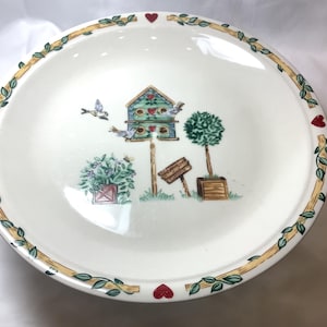 Puede incluir: Un plato de cerámica blanco con un borde verde y amarillo y corazones rojos. El centro del plato presenta una casa para pájaros con un techo verde y detalles azules, rodeado de pájaros, árboles y flores.