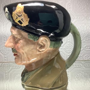 May include: A ceramic jug shaped like a man's head, sporting a black beret with a gold emblem. The jug features a light green handle and is painted in realistic colours.