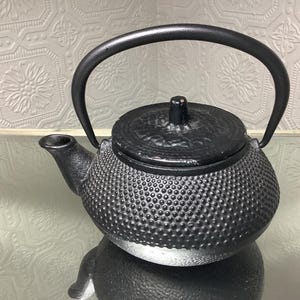 May include: Black cast iron teapot with a textured, dimpled surface. The teapot has a curved handle and a lid with a small knob. The spout is short and curved. The teapot is designed for brewing tea.
