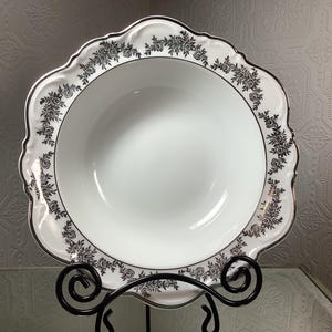 May include: White ceramic serving bowl with a scalloped edge and a silver floral design. The bowl is displayed on a black metal stand. A silver trim accents the rim of the bowl.