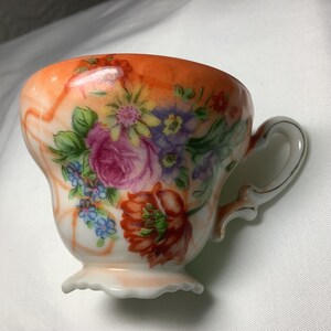 Peut inclure: Une tasse à thé blanche avec un motif floral. La tasse a un bord festonné et une anse. Le motif floral présente des fleurs roses, jaunes, orange et bleues sur un fond blanc avec une bordure de couleur pêche.