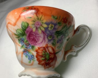 May include: A white teacup with a floral design. The cup has a scalloped edge and a handle. The floral design features pink, yellow, orange, and blue flowers on a white background with a peach colored rim.