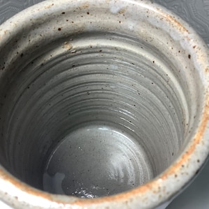 Puede incluir: Un recipiente cil&iacute;ndrico de cer&aacute;mica con un esmalte gris claro moteado y anillos horizontales visibles en las paredes interiores. El borde presenta un tono marr&oacute;n anaranjado tostado y la superficie tiene peque&ntilde;as manchas de color marr&oacute;n oscuro.