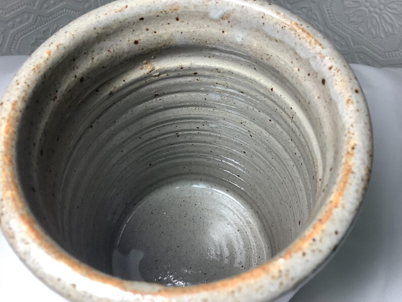 Puede incluir: Un recipiente cil&iacute;ndrico de cer&aacute;mica con un esmalte gris claro moteado y anillos horizontales visibles en las paredes interiores. El borde presenta un tono marr&oacute;n anaranjado tostado y la superficie tiene peque&ntilde;as manchas de color marr&oacute;n oscuro.