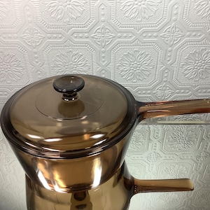 Peut inclure: Une casserole en verre ambré translucide avec un couvercle assorti et un long manche. L'ustensile de cuisine présente une surface lisse et réfléchissante ainsi qu'un bouton circulaire au centre du couvercle, avec une teinte brune profonde.