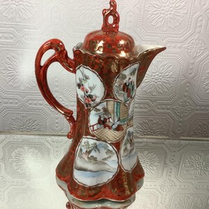 May include: An antique ceramic coffee pot in red and gold. The pot has a lid and handle, and is decorated with hand-painted scenes in circular frames. The handle and lid are red, while the body has a textured pattern with gold accents.