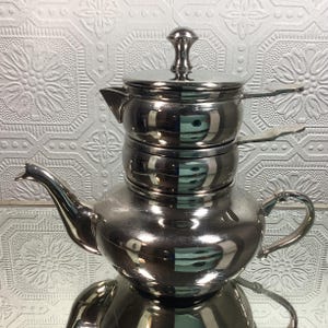 May include: A shiny, stainless steel tea set with a teapot, two stacked pots, and a lid. The teapot has a curved spout and a handle. The set is reflective, with the background visible in its surface.