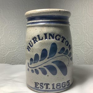 May include: A stoneware crock with a beige speckled finish. The word "BURLINGTON" is arched across the top, with a blue floral design below. The text "EST. 1865" is at the bottom. The rim and top band are blue.