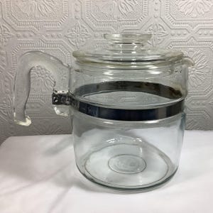 May include: A clear glass coffee carafe featuring a wide, dark metal band encircling the upper body. The carafe includes a matching clear glass lid with a central knob handle and a sturdy, integrated glass handle on the side for pouring.