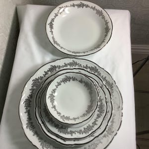 May include: A collection of white porcelain dinnerware with a silver floral design around the rims. The set includes various sized plates and bowls, perfect for serving a meal. The plates have a scalloped edge.