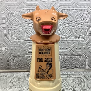 Könnte beinhalten: Ein beigefarbener und cremefarbener Milchkännchen aus Kunststoff in Form eines Kuhkopfes mit rotem Mund. Die Vorderseite zeigt ein rechteckiges Etikett mit dem Text MOO-COW CREAMER FOR SALE und einer Abbildung einer Person mit Tablett und dem Text ask your waitress!