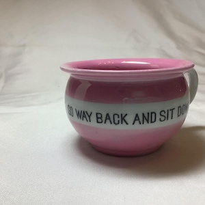 May include: A pink ceramic bowl with a white stripe and the text "GO WAY BACK AND SIT DOWN" in black. The bowl has a small handle and a rounded shape, suitable for decorative use.
