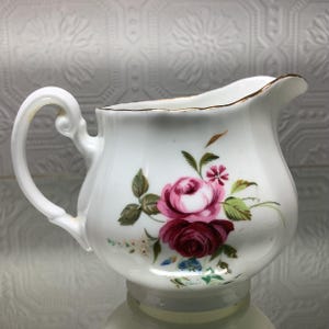 Peut inclure: Crémier en porcelaine blanche avec un motif floral de roses dans les tons de rose et de bordeaux. Le crémier a une anse incurvée et une bordure dorée sur le bord. Le motif floral comprend des feuilles vertes et de petites fleurs bleues et roses.