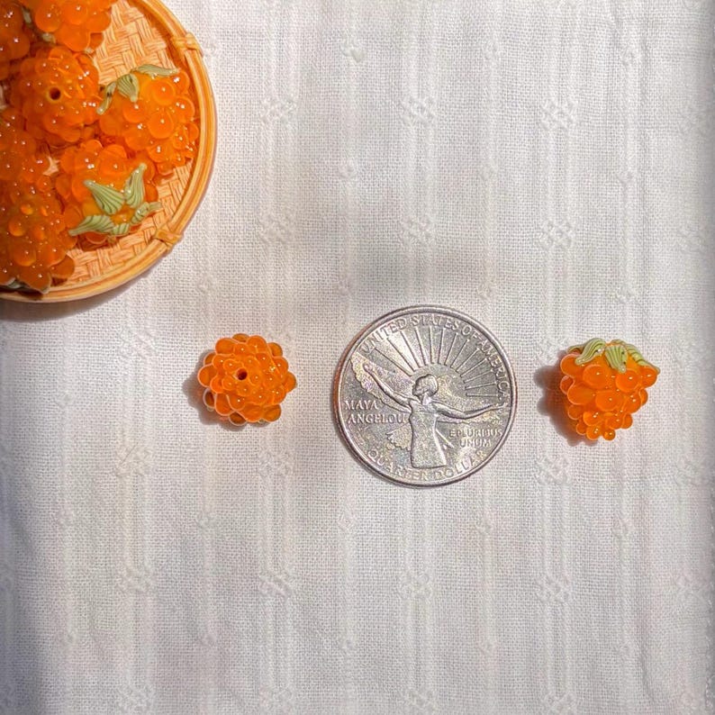 May include: Orange berry-shaped beads with small green leaves, shown next to a US quarter coin for scale. The beads have a textured, bumpy surface resembling real fruit. A small collection of these beads is contained in a round woven basket.