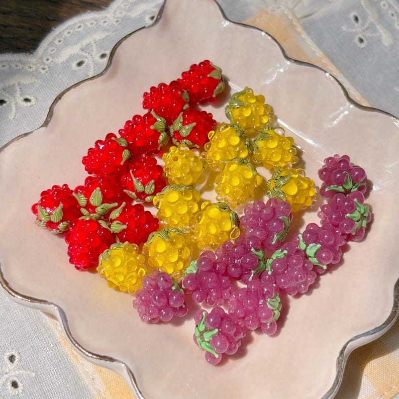 May include: A collection of small, translucent berry-shaped beads in red, yellow, and purple. Each bead features a textured, bumpy surface resembling a raspberry or blackberry, accented with a small, painted green leaf detail at the top.