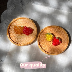 May include: Two small, round, woven bamboo baskets, each containing a pair of miniature fruit-shaped charms. One basket holds an orange and a red berry charm, while the other holds a yellow and a red berry charm. Text reads: Our quality.